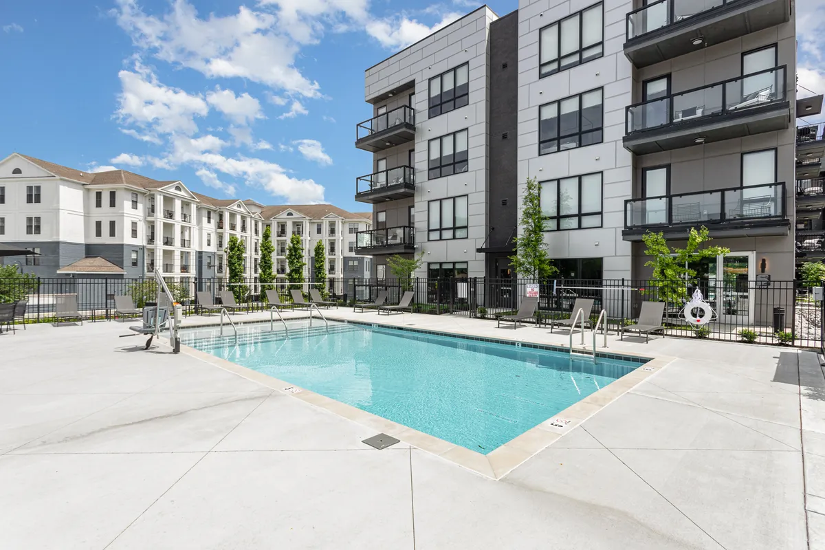 An inviting exterior shot of a resort-style swimming pool surrounded by apartment balconies and lush green trees. The area is perfect for swimming, sunbathing, and socializing, with the soft light of sunset creating a relaxed atmosphere.
