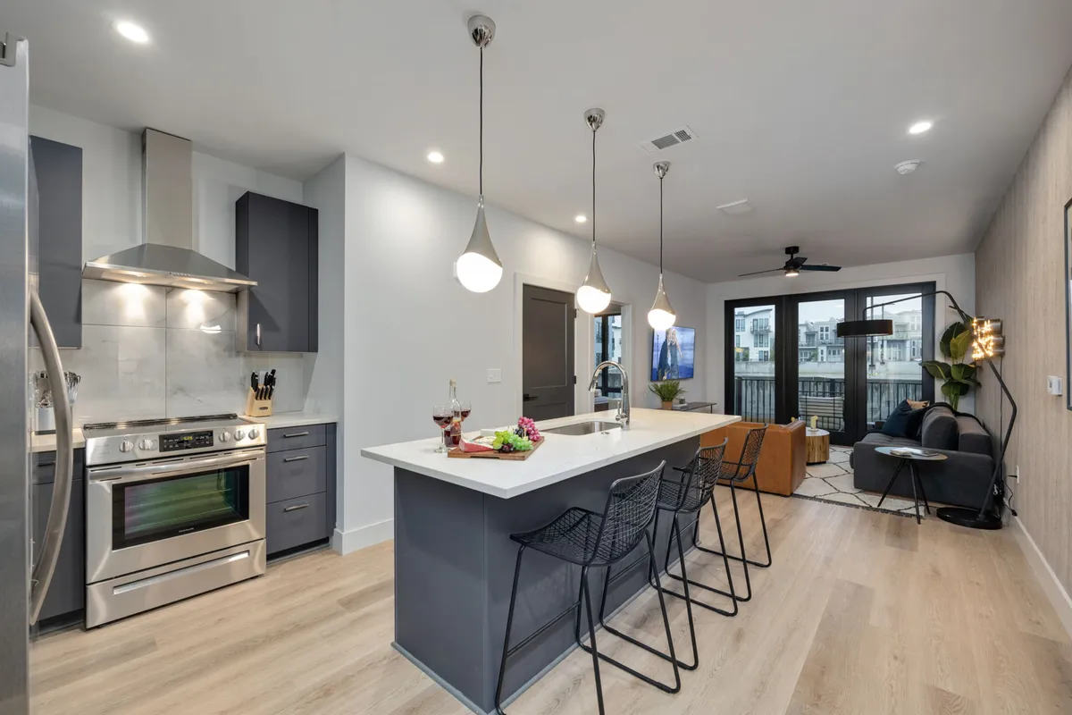 A contemporary, open-concept kitchen with a central island, stainless steel appliances, stylish seating, and an open balcony door visible in the background. The scene suggests a seamless transition from cooking to entertaining, with the balcony inviting in light and city views.