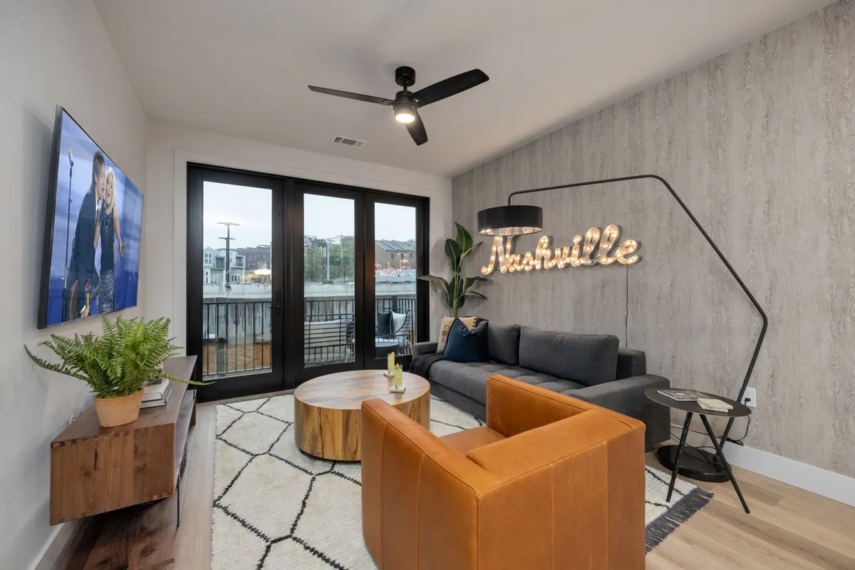 The image showcases a cozy, stylish living room featuring a plush sofa and coffee table, a prominent 'Nashville' sign adding local flair, and a large television—creating an inviting space for relaxing, entertaining, or enjoying a night in. Natural light suggests easy access to a balcony for sunset views, completing the welcoming ambiance.