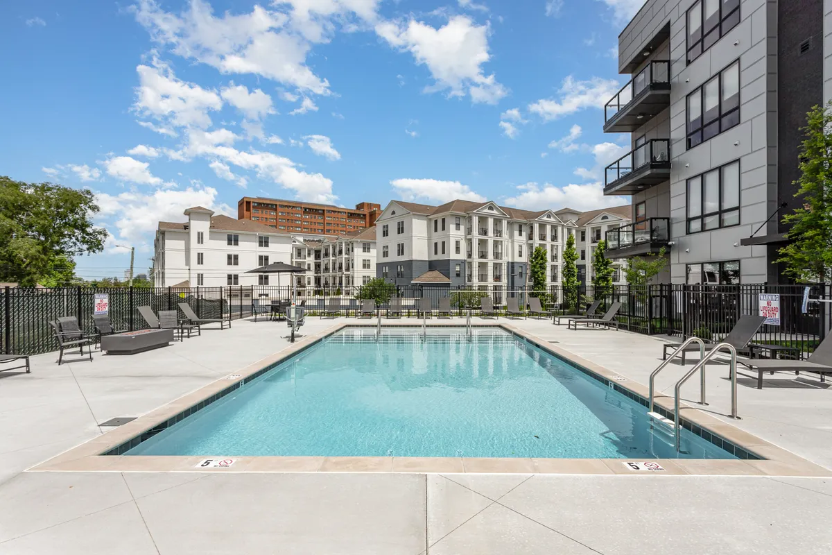 This image shows a sparkling swimming pool surrounded by a modern lounge area and apartment buildings. The setting is active and inviting, with hints of sunbathing, swimming, and socializing by the poolside. The pool area is designed for both relaxation and community gatherings, evoking the feel of a resort.