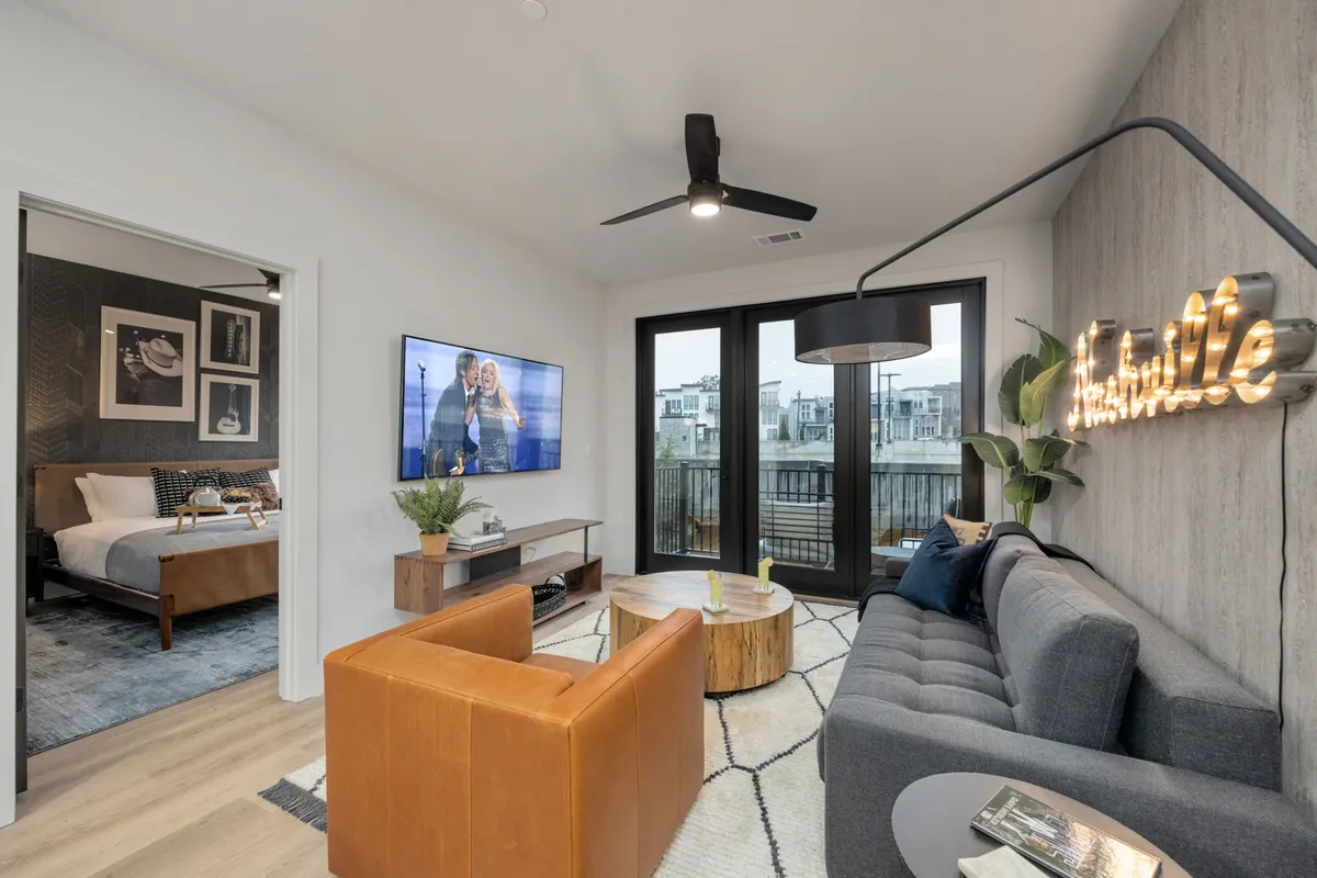 A contemporary living room with a plush sofa, large windows that let in abundant natural light, a stylish wooden coffee table, a Smart TV, and a Nashville neon sign on the wall. The space feels open, bright, and welcoming, perfect for social gatherings or relaxation.