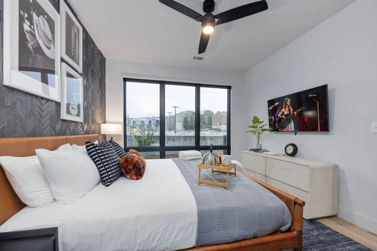 A bedroom with a comfortable bed, window views, a Smart TV mounted opposite the bed, and decorative items on the dresser. The mood is inviting and comfortable, ideal for unwinding with a movie or enjoying a quiet morning in bed.